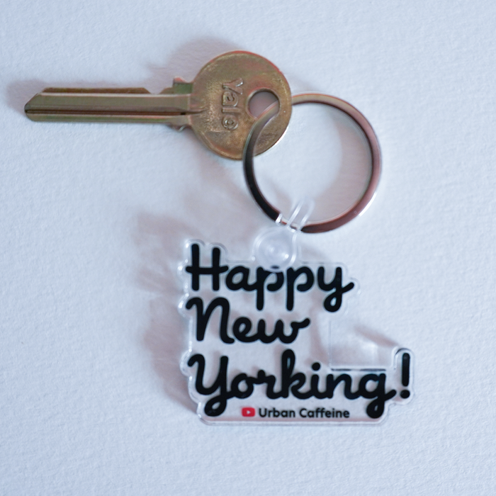 A keychain with an acrylic charm and a key attached. The charm design has the words "Happy New Yorking" in fun cursive typeface with the words "Urban Caffeine" underneath it.