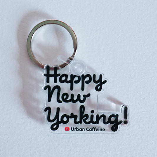 A clear acrylic keychain with the phrase "Happy New Yorking!" in black stylized text, attached to a metal key ring. Below the text is a small red Youtube logo followed by "Urban Caffeine." The keychain is laid on a white surface and has a custom shape that outlines the lettering.