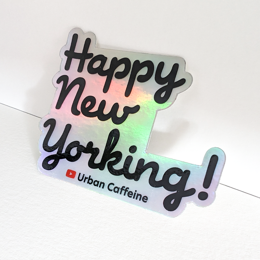 A holographic sticker with the phrase "Happy New Yorking!" in black stylized text, affixed to a white surface at an angle. Below the phrase is a red Youtube logo followed by the words "Urban Caffeine." The sticker has a die-cut border following the shape of the letters.