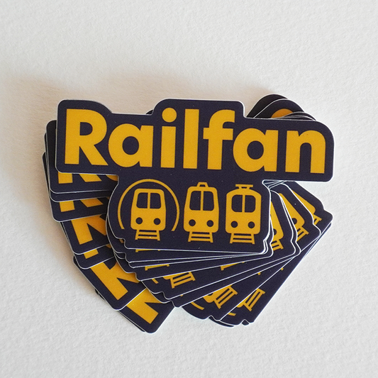 A stack of dark vinyl stickers with the word "Railfan" printed in bold yellow letters. The design includes three stylized yellow trains below the text, on tracks that are implied by a series of horizontal yellow lines. The stickers have a die-cut shape around the contour of the text and trains.